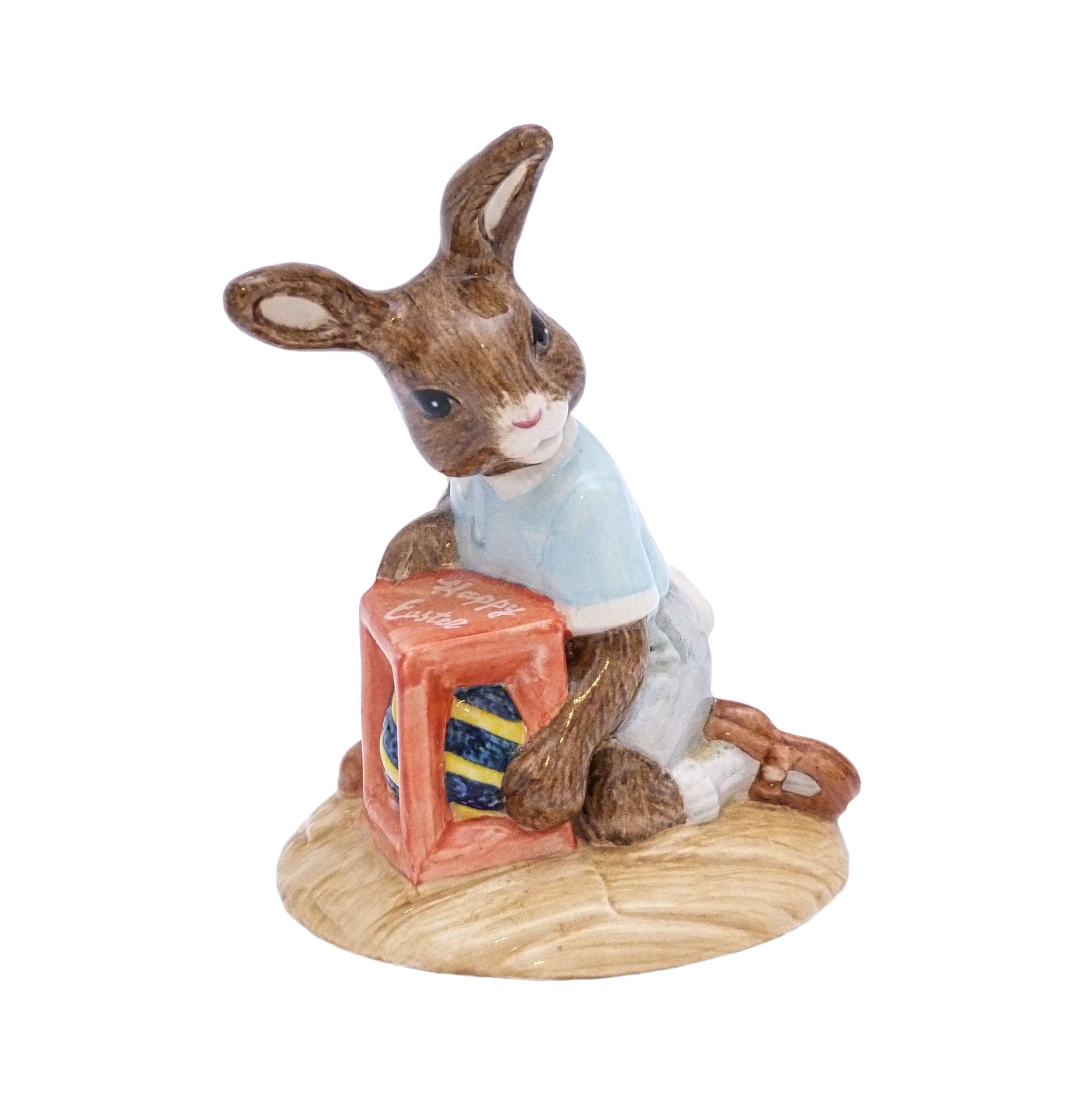 The bunny is kneeling holding his large Easter egg wrapped in blue and gold, the egg is in a red box with Happy Easter written on the top.