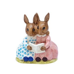 The two bunnykins are sitting holding a book. One is wearing a pink dress the other a white dress with blue spots.