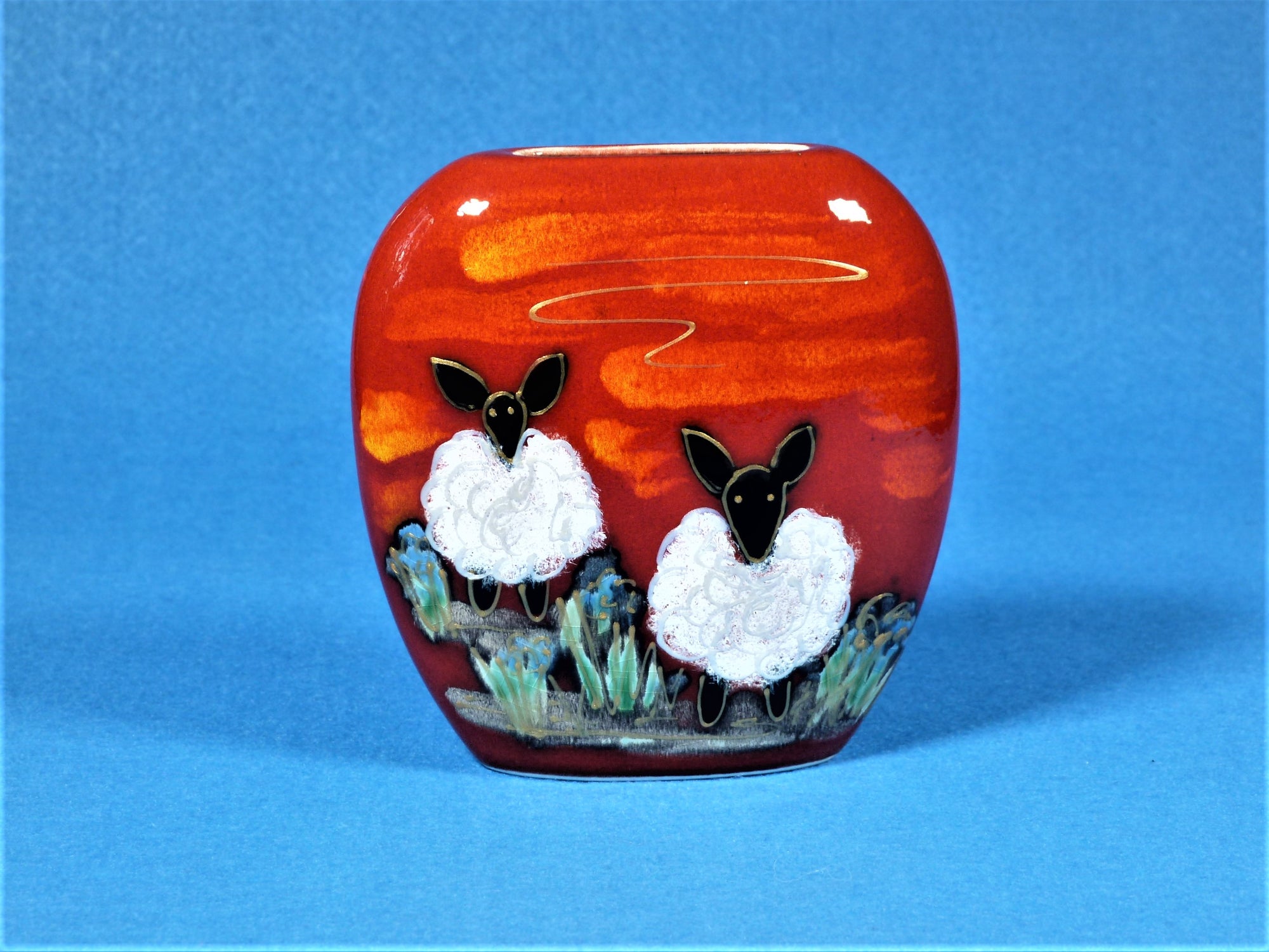 A small red vase with two white sheep with black heads on the front and one on the back.